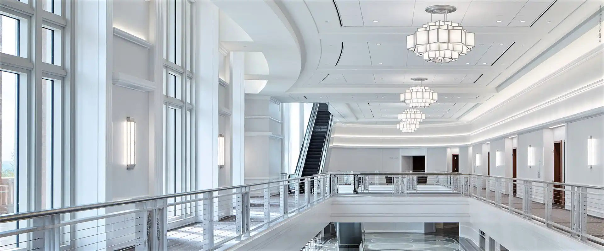 A clean, modern, multi-story interior space with a wide hallway, high ceilings, large windows, an escalator, and decorative lighting.