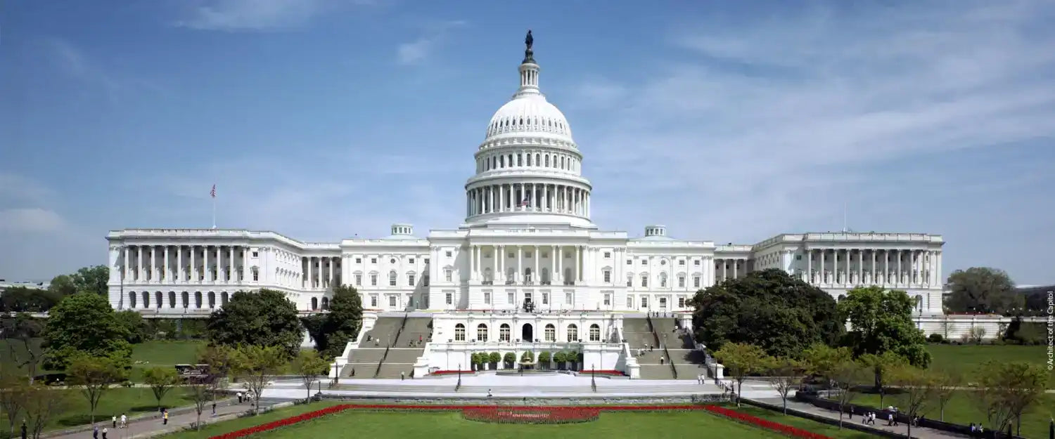 The United States Capitol Building, a white neoclassical structure with a large dome, columns, and grand staircases, surrounded by green lawns.