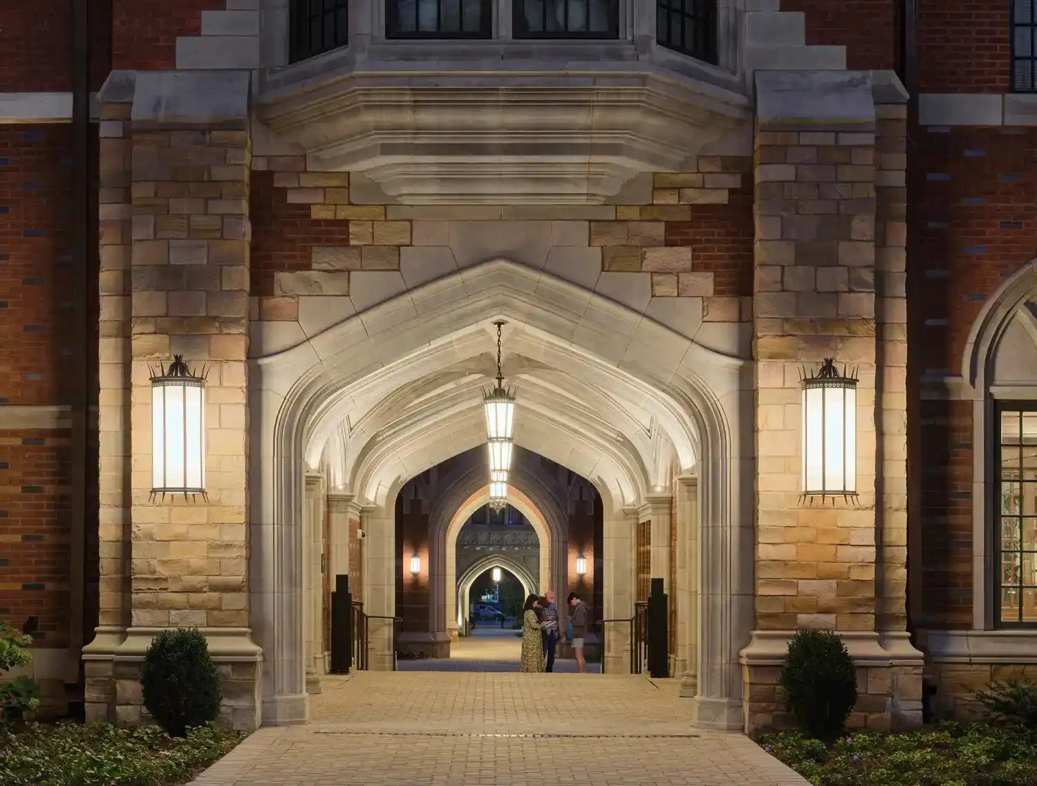 An illuminated stone archway leading to a series of connected arches, with decorative hanging lanterns and wall-mounted lights.