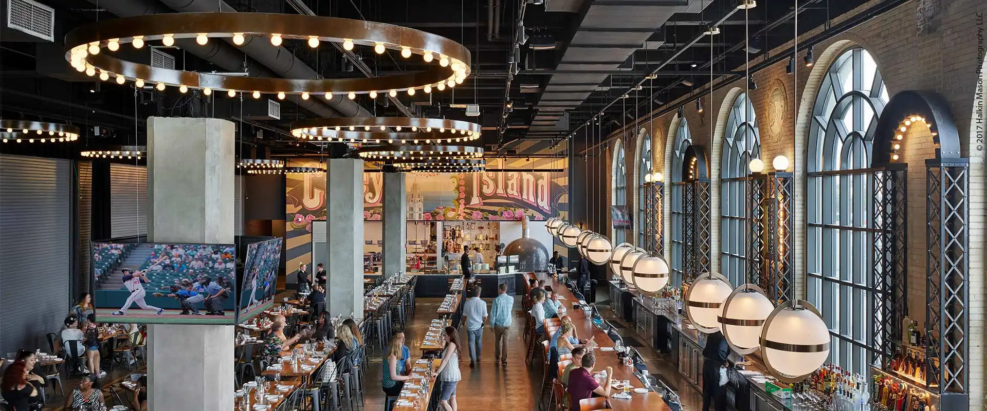 A spacious, modern restaurant or bar with high ceilings, long communal tables, arched windows, and decorative lighting fixtures.