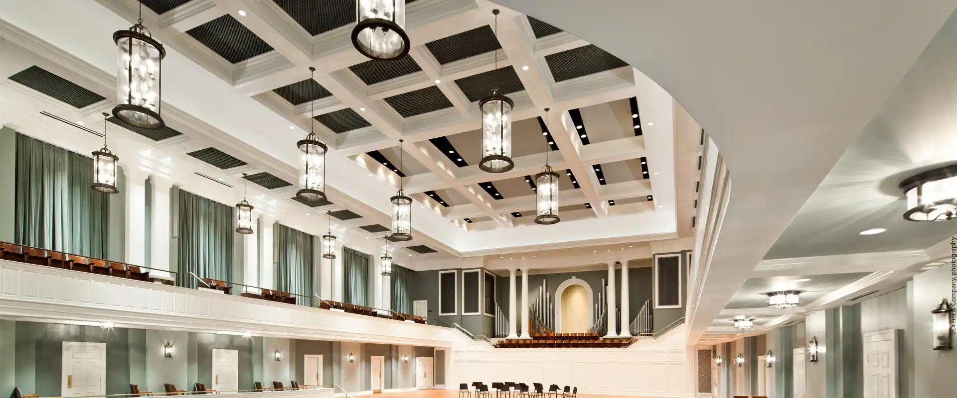 A grand hall with a high, coffered ceiling, elegant hanging lanterns, a balcony, and a stage with an arched alcove.