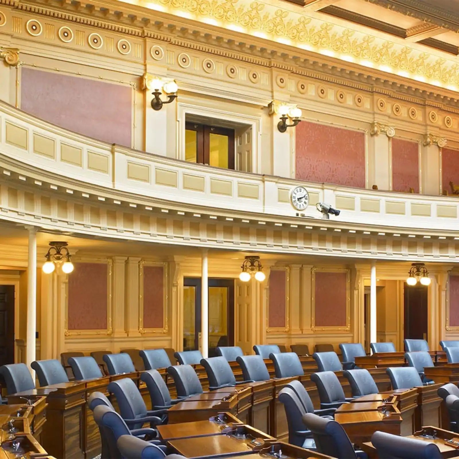 An ornate, multi-tiered assembly hall with rows of dark wood desks and blue upholstered chairs, featuring a balcony, decorative walls, and ceiling, with several wall-mounted lamps and a clock.